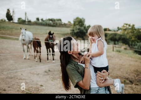 Madre sorridente che porta la figlia allegra mentre si trova contro i cavalli terra Foto Stock