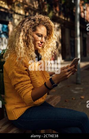 Giovane donna con lunghi capelli ricci biondi con smartphone mentre si è seduti in città Foto Stock