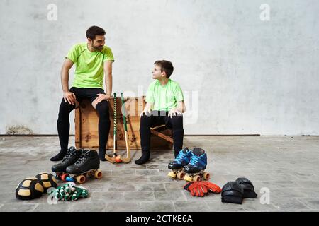 Padre e figlio che parlano mentre si siedono su una scatola di legno vicino attrezzature sportive contro il muro in campo Foto Stock