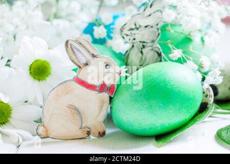 Uova di Pasqua dipinte verdi e blu; conigli dipinti, fiori bianchi su sfondo bianco di legno, fuoco selettivo Foto Stock