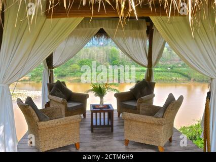 Un ambiente tropicale di comode sedie in rattan su un gazebo aperto, con tende di lino bianco, che si affaccia su un ambiente rurale. Foto Stock