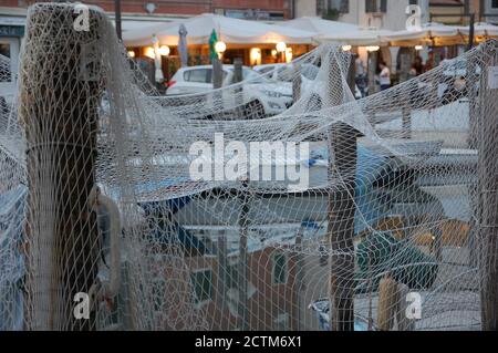 una rete di pesce che asciuga su alcuni pali di legno in un canale di Chioggia nella laguna veneta con barche intorno e luci di strada sera sullo sfondo Foto Stock