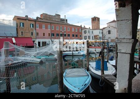 una rete di pesce che asciuga su alcuni pali di legno in un canale di Chioggia nella laguna veneta con barche intorno e luci di strada sera sullo sfondo Foto Stock