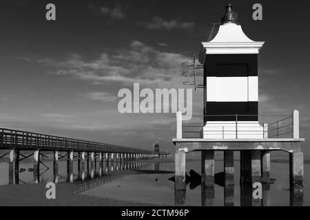 Immagine in bianco e nero di un faro e di un molo in legno sotto un cielo drammatico. Lignano Sabbiadoro, Friuli Venezia Foto Stock