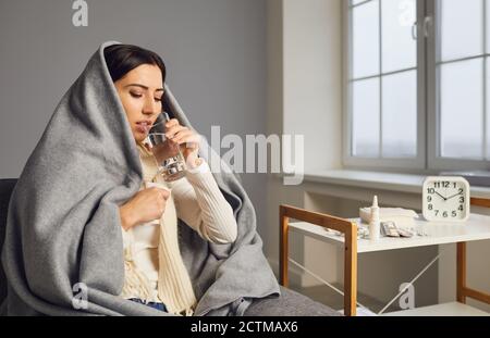 Vista laterale della giovane donna in coperta ha preso freddo e prendendo medicina di influenza, acqua potabile. Foto Stock