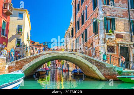 Venezia, Italia, 13 settembre 2019: Ponte attraverso lo stretto canale d'acqua con barche ormeggiate tra vecchi edifici colorati con balconi e pareti, cielo blu, Regione Veneto. Tipico paesaggio urbano veneziano Foto Stock