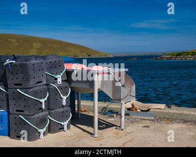 In una giornata di sole in estate, pentole di aragosta di plastica nera accatastate su un molo pronto per l'uso, accanto a un tavolo di eviscerazione del pesce - preso a Shetland, Scozia, Regno Unito Foto Stock