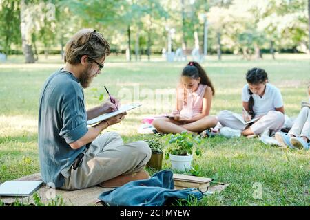 Insegnante con bearded concentrato in bicchieri seduti con gambe incrociate e. prendere appunti negli appunti mentre gli studenti eseguono attività nel parco Foto Stock