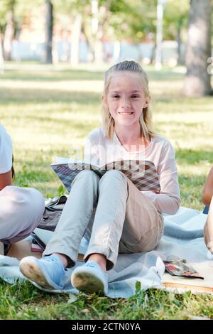 Ritratto di sorridente bella ragazza bionda seduta su coperta dentro parcheggiare e prendere appunti nel libro degli esercizi mentre si svolge un'attività scolastica Foto Stock