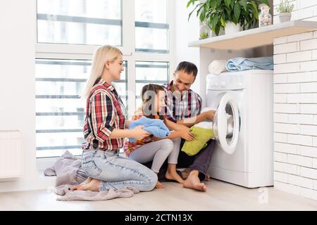 madre di famiglia, padre e bambina piccola helper in lavanderia vicino lavatrice e vestiti sporchi Foto Stock