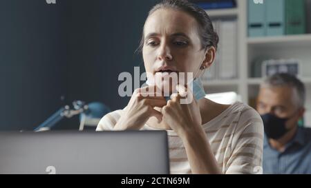 Donna che lavora in ufficio e ha difficoltà a respirare con la maschera facciale, sta tirando la maschera verso il basso Foto Stock