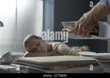 Stanca donna pigra che dormiva sulla scrivania dell'ufficio, il suo capo porta un mucchio di documenti alla sua scrivania Foto Stock