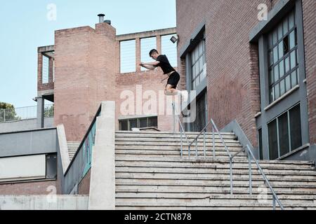 Vista laterale di un giovane maschio concentrato che salta sopra i gradini di pietra in città mentre facendo parkour e mostrando trucco Foto Stock