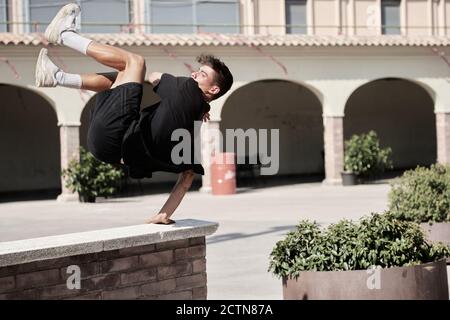 Vista laterale del maschio concentrato che salta sopra la recinzione di pietra e. equilibrio sul braccio durante l'esecuzione di acrobazie e fare parkour Foto Stock