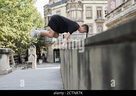 Vista laterale del maschio concentrato che salta sopra la recinzione di pietra e. equilibrio sul braccio durante l'esecuzione di acrobazie e fare parkour Foto Stock