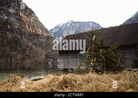 Casa di legno solitario situata sulla riva del lago contro le montagne rocciose coperto di alberi e neve in autunno giorno Foto Stock