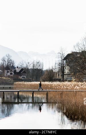 Vista laterale di una persona irriconoscibile che cammina sul lungomare di legno lago calmo con riva erbosa in piccolo insediamento situato in terreno montagnoso in autunno giorno Foto Stock
