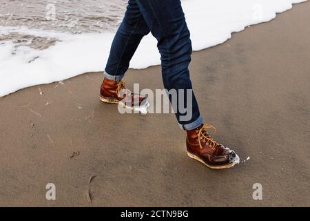 Alto angolo di raccolto anonimo maschio che cammina su sabbia bagnata spiaggia vicino al mare schiumoso Foto Stock