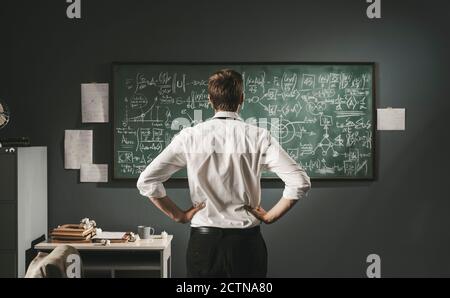 Matematico rivedere le formule matematiche sulla lavagna, sta pensando e scoprendo nuove teorie, vista posteriore Foto Stock