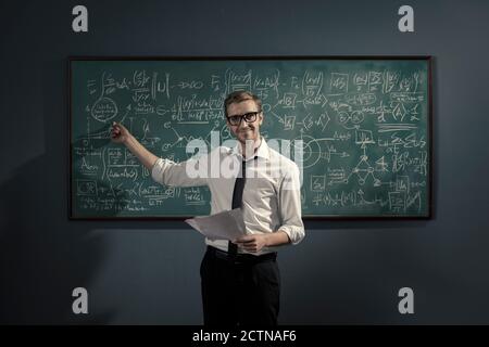 Un giovane e sicuro professore di matematica che si trova di fronte alla lavagna e spiega le formule, sta indicando e sorridendo Foto Stock