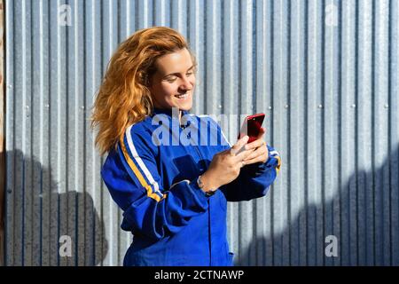 Donna molto felice in uniforme e occhiali protettivi in piedi vicino costruzione di metallo e telefono cellulare di navigazione mentre hanno la rottura durante il lavoro Foto Stock