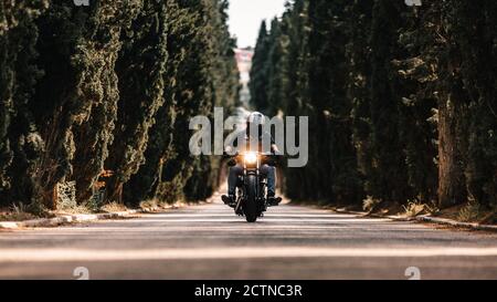 Motociclista in giacche di pelle nera e casco che cavalcano moto potente su strada asfaltata che conduce tra verde foresta in campagna Foto Stock