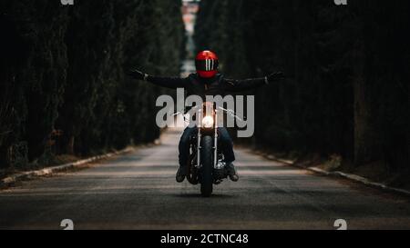 Biker con giacche in pelle nera e casco a braccia aperte guida potente moto su strada asfaltata che conduce tra foresta verde in campagna Foto Stock