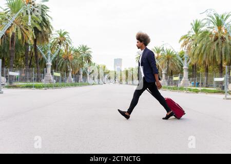 Vista laterale di un giovane uomo nero in tuta trascinando una valigia con ruote lungo la strada su una linea alberata viale Foto Stock