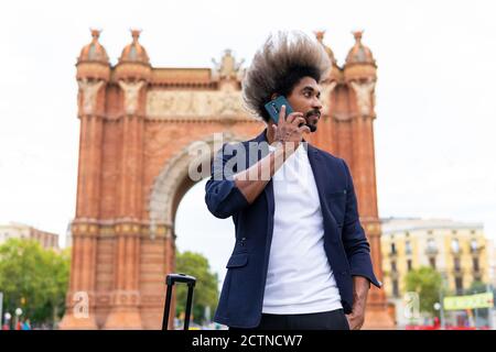 Giovane uomo nero in tuta e valigia in piedi per strada parlando con il suo cellulare Foto Stock