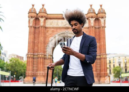 Giovane uomo nero in tuta e valigia in piedi per strada parlando con il suo cellulare Foto Stock
