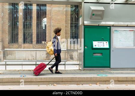 Vista laterale dell'uomo nero con i capelli afro che camminano sopra una stazione del tram con una valigia su ruote e un borsa appesa alla spalla Foto Stock