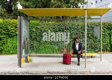 Uomo nero con capelli afro e una valigia seduta a. una fermata dei trasporti pubblici mentre parla con il suo cellulare Foto Stock