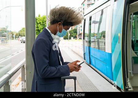 Uomo nero con capelli afro e una maschera viso utilizzando il suo telefono cellulare di fronte al tram con un valigia accanto a lui e un auricolare wireless intorno a lui collo Foto Stock