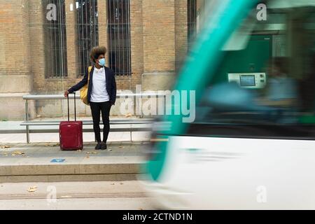 Uomo nero con capelli afro e maschera protettiva viso in piedi sulla strada con una valigia e una borsa appesa dalla sua spalla di fronte a un tram che è in movimento Foto Stock