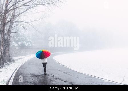 Vista posteriore di persona anonima sotto ombrello colorato che cammina lungo Strada nel parco invernale in giornata innevata nei Pirenei Foto Stock