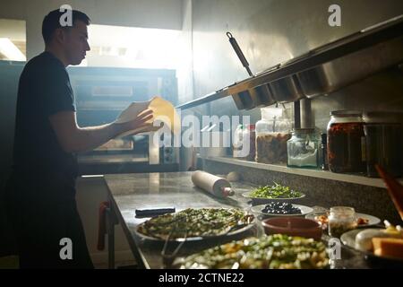 Vista laterale dell'uomo che si capovolgerà e si affetta l'impasto sul tavolo con farina mentre stretching pizza base in cucina ristorante Foto Stock