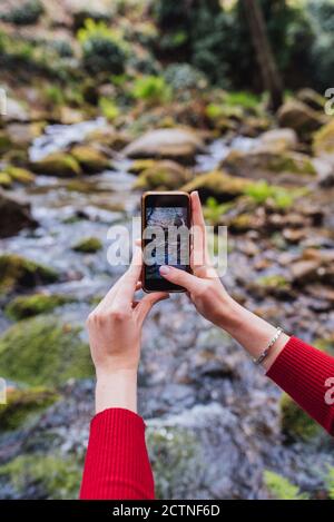 Vista posteriore di anonimo turista femminile seduto sulla riva di fiume in boschi e scattare foto di paesaggio incredibile in Valle del Jerte Foto Stock