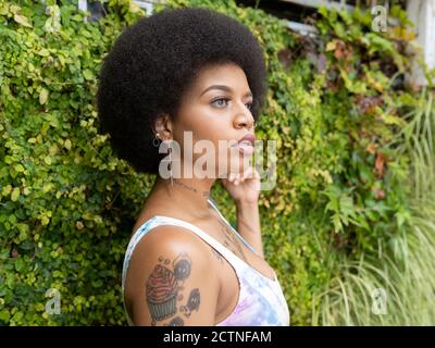 Vista laterale dell'elegante donna afroamericana con tatuaggi e piercing in piedi nel parco urbano in estate e guardando lontano Foto Stock