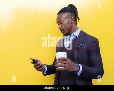 Imprenditore maschile nero in abito elegante in piedi con caffè da asporto in tazza di carta e cellulare guardando via il giallo sfondo Foto Stock