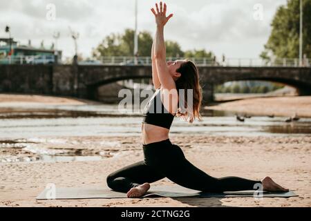 Vista laterale della donna serena in reggiseno e leggings seduti Su tappetino yoga in Ardha Kapotasana con braccia sollevate mentre guardare in su e mettere in pratica la consapevolezza Foto Stock