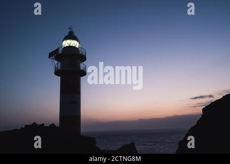 Basso angolo di spettacolare scenario di faro illuminato situato su Collina rocciosa vicino al mare contro il cielo del tramonto a Tenerife Foto Stock
