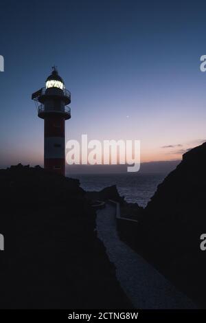 Basso angolo di spettacolare scenario di faro illuminato situato su Collina rocciosa vicino al mare contro il cielo del tramonto a Tenerife Foto Stock