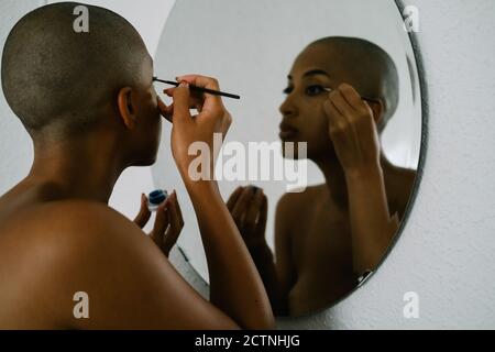 Vista laterale della donna afroamericana con testa calva che fa trucco e applicazione eyeliner liquido con spazzola mentre si guarda in speculare Foto Stock