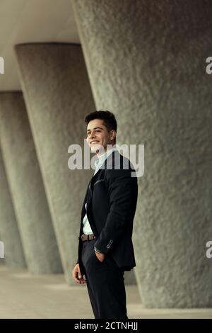 Vista laterale del soddisfare handsome uomo d'affari in piedi con la mano dentro tasca vicino all'edificio in città e guardare lontano Foto Stock