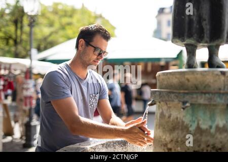 Giovane uomo casual cucasual lavarsi le mani con acqua dal pubblico fontana della città in una calda giornata estiva Foto Stock