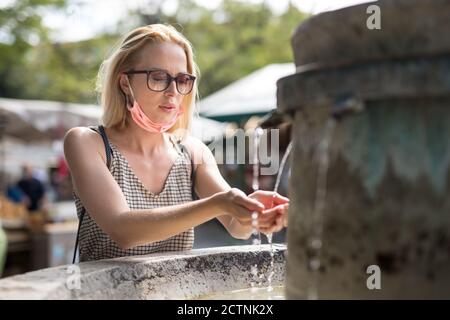 Assetata giovane donna casual cucasual con maschera medica viso bere acqua dalla fontana della città pubblica in una calda giornata estiva. Nuova vita durante il covid Foto Stock
