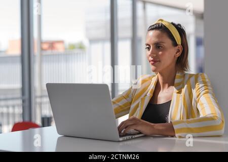 Donna in stato di gravidanza lavoratrice a distanza positiva in elegante giacca a righe e archetto seduto al tavolo e guardando il computer portatile mentre si lavora su progetto a casa Foto Stock