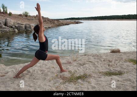 Vista laterale del corpo della femmina sottile in abbigliamento sportivo Asana guerriero con le braccia sollevate durante lo yoga vicino al lago Foto Stock