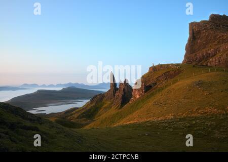 Paesaggio soleggiato con le alte scogliere che si innalzano sul lago e il mare al mattino e piccole figure di persone. L'Old Man of Storr, l'isola di Skye, Scozia, Regno Unito Foto Stock