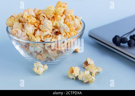 Una ciotola trasparente di popcorn su una superficie blu. Accanto a un computer portatile e alle cuffie. La famiglia che guarda i film. Spazio di copia. Foto Stock